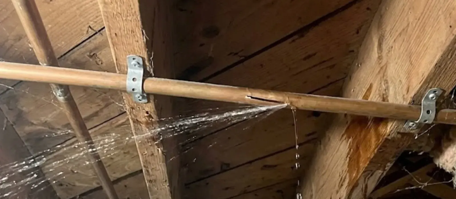 Split copper water pipe spraying water between wooden ceiling joists
